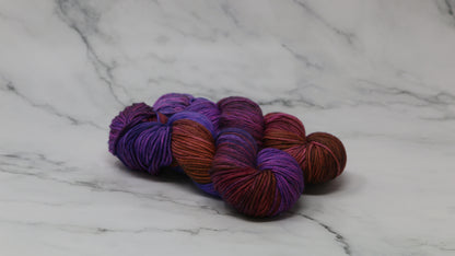 A couple of variegated yarn skeins in purple, burgundy, and orange tones, arranged on a white marble surface with a matching marble background.