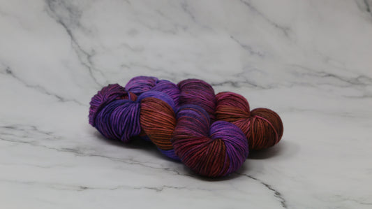 A couple of variegated yarn skeins in purple, burgundy, and orange tones, arranged on a white marble surface with a matching marble background.