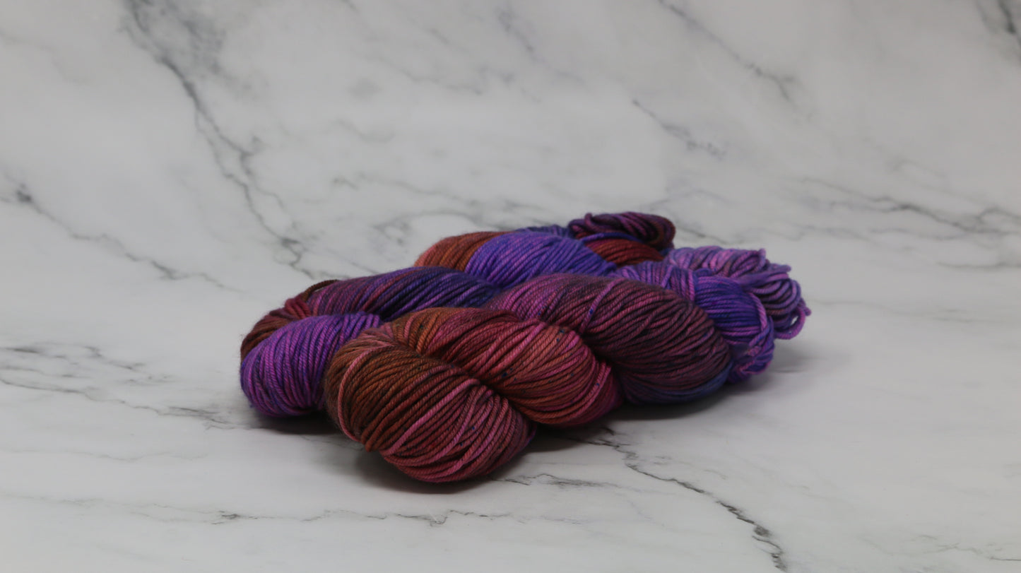 A couple of variegated yarn skeins in purple, burgundy, and orange tones, arranged on a white marble surface with a matching marble background.