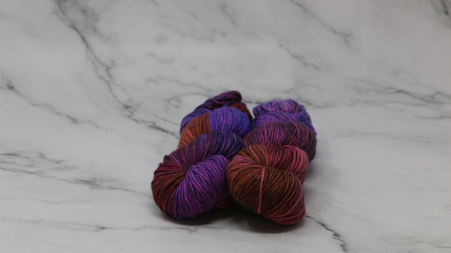 A couple of variegated yarn skeins in purple, burgundy, and orange tones, arranged on a white marble surface with a matching marble background.