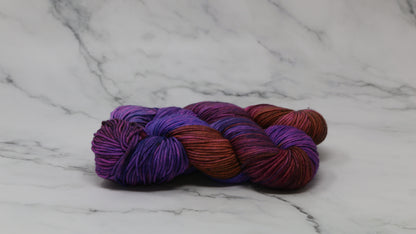 A couple of variegated yarn skeins in purple, burgundy, and orange tones, arranged on a white marble surface with a matching marble background.