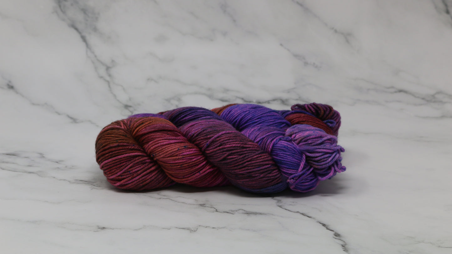 A couple of variegated yarn skeins in purple, burgundy, and orange tones, arranged on a white marble surface with a matching marble background.
