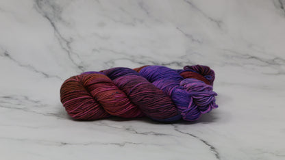 A couple of variegated yarn skeins in purple, burgundy, and orange tones, arranged on a white marble surface with a matching marble background.