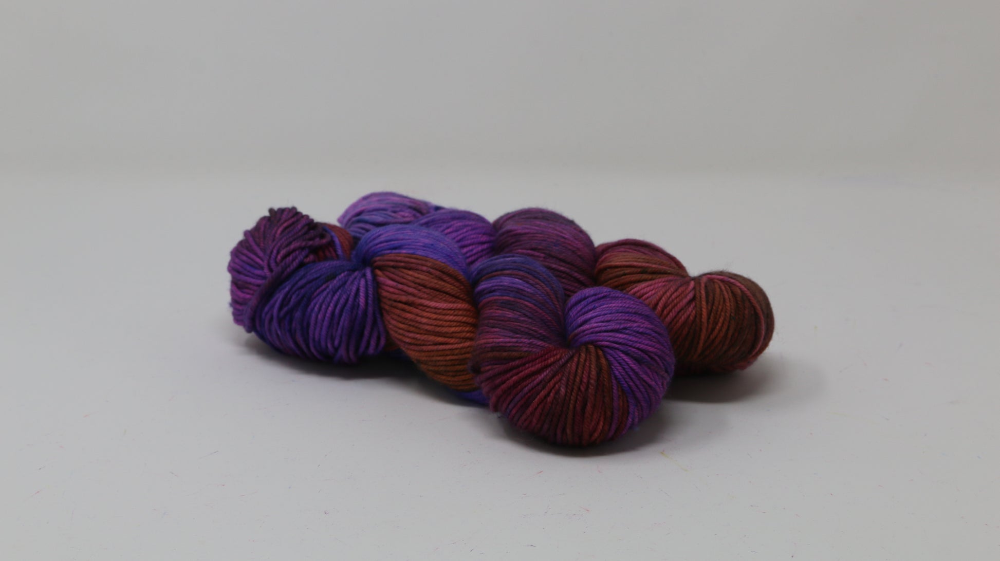 A couple of variegated yarn skeins in purple, burgundy, and orange tones, arranged on a white surface with a matching white background.