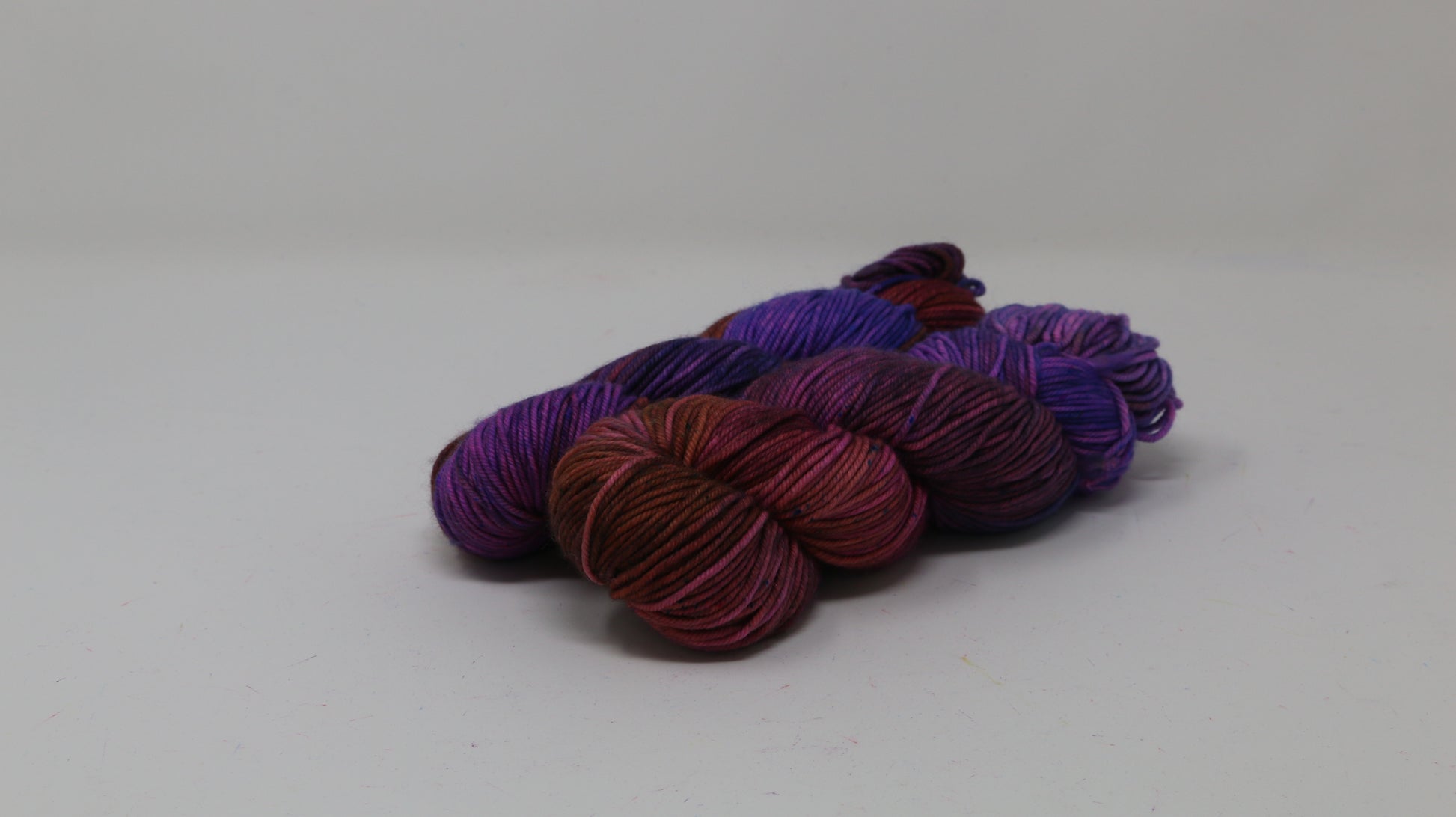 A couple of variegated yarn skeins in purple, burgundy, and orange tones, arranged on a white surface with a matching white background.
