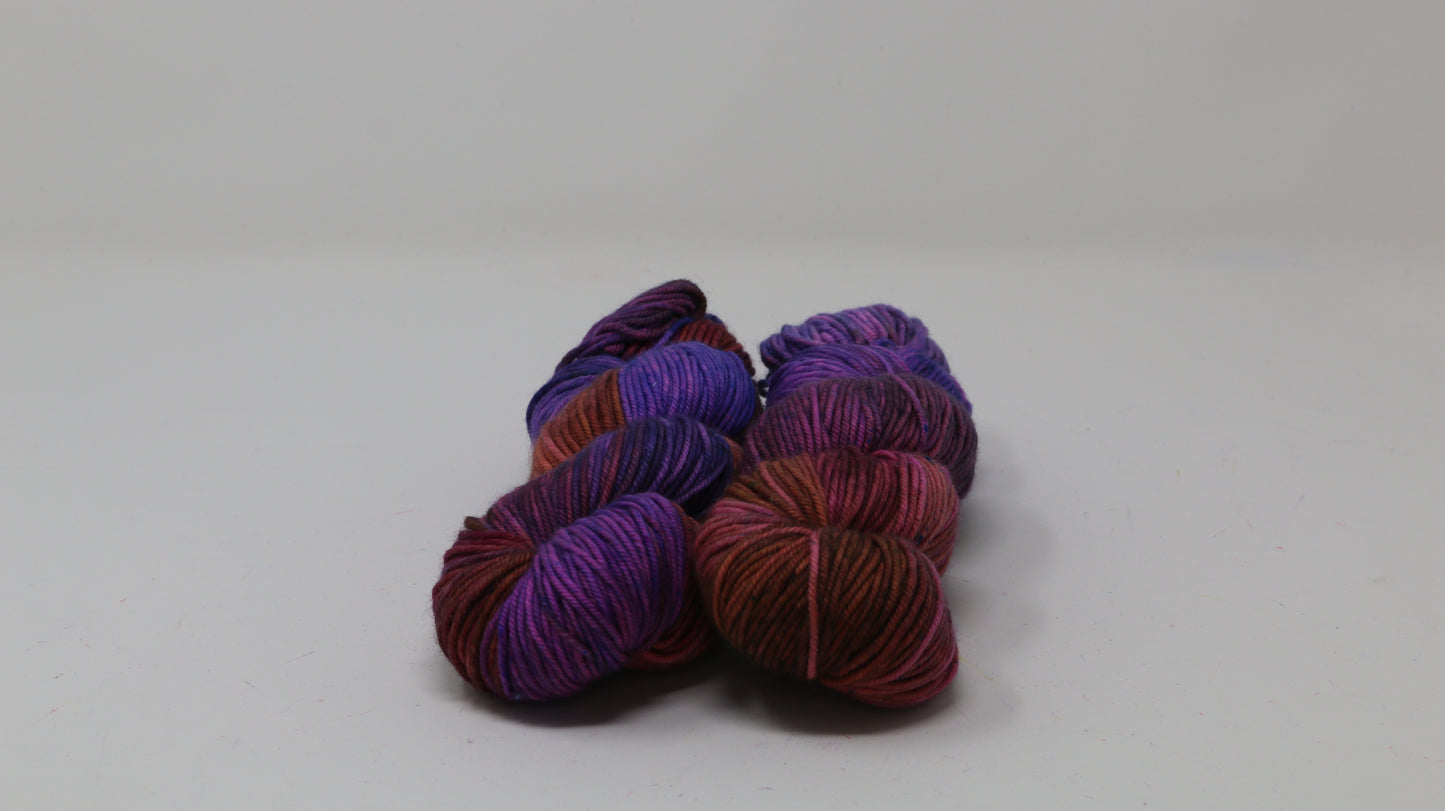 A couple of variegated yarn skeins in purple, burgundy, and orange tones, arranged on a white surface with a matching white background.
