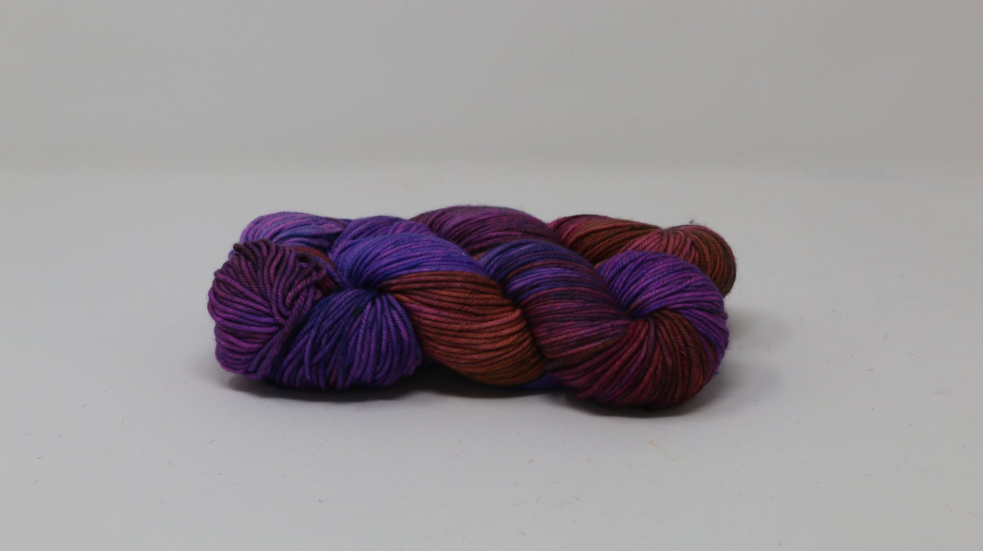 A couple of variegated yarn skeins in purple, burgundy, and orange tones, arranged on a white surface with a matching white background.