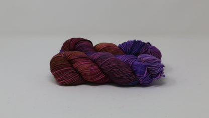 A couple of variegated yarn skeins in purple, burgundy, and orange tones, arranged on a white surface with a matching white background.