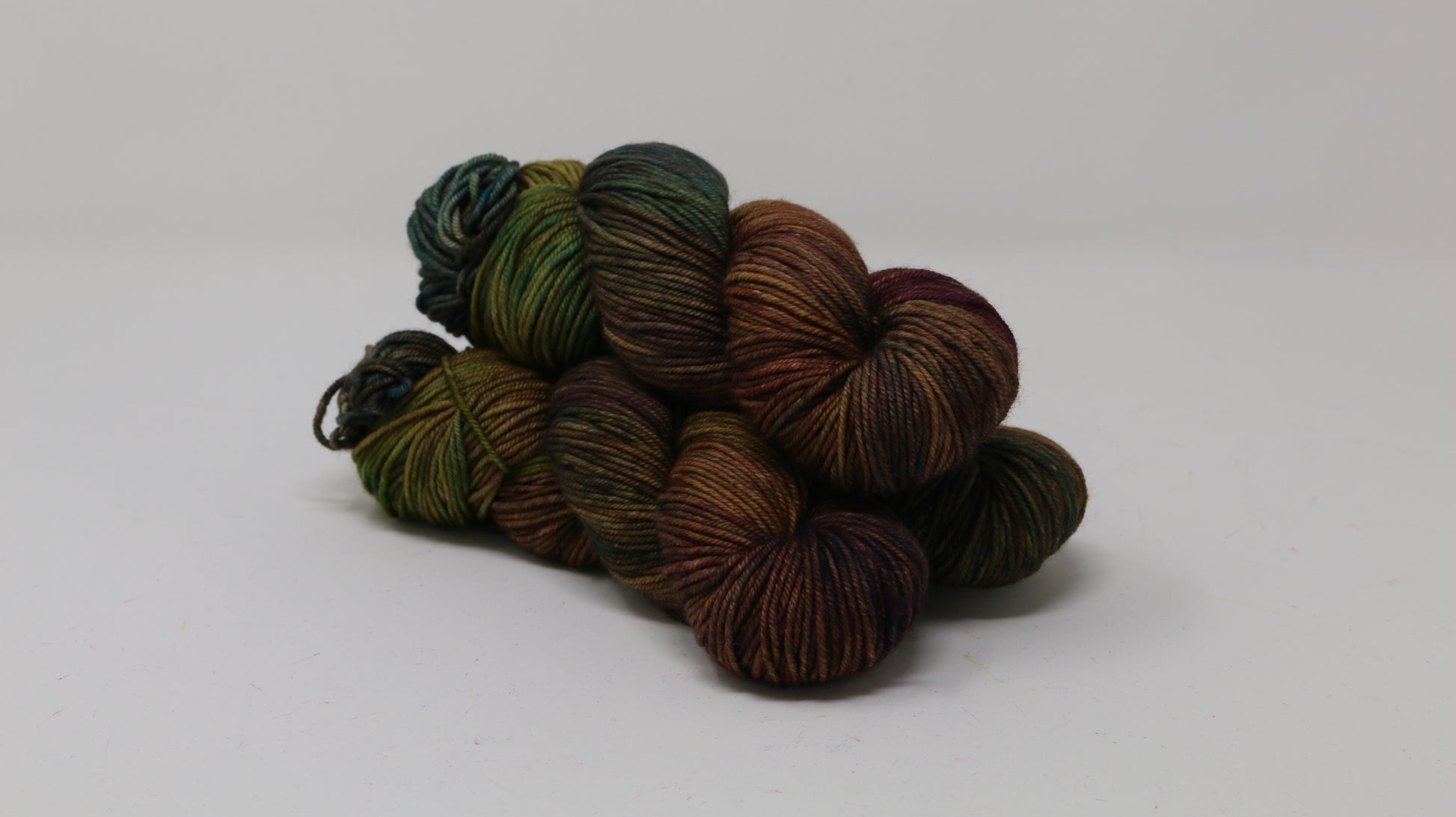3 skeins of hand-dyed yarn in earthy, variegated shades of green, brown, and burgundy, displayed on a white surface and background.