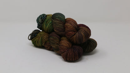 3 skeins of hand-dyed yarn in earthy, variegated shades of green, brown, and burgundy, displayed on a white surface and background.