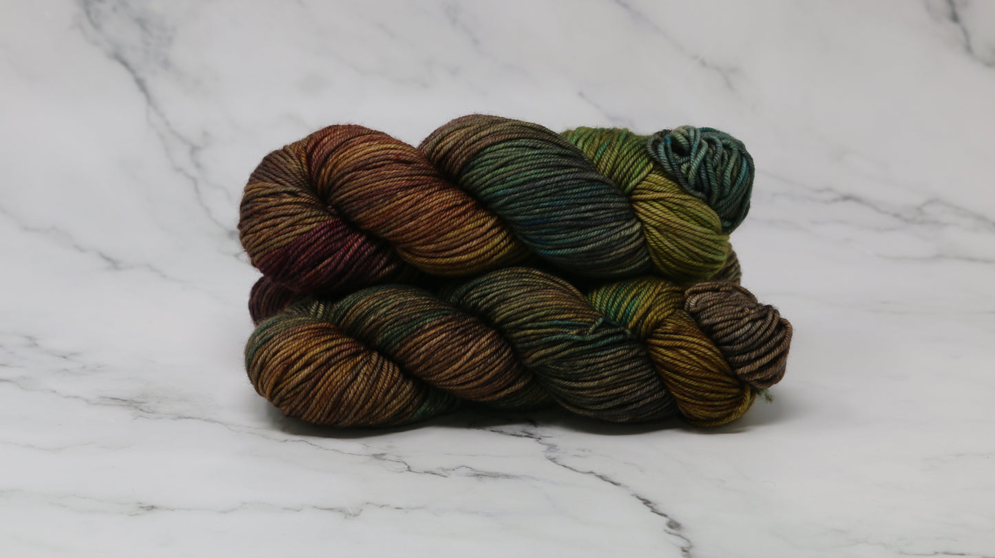 3 skeins of hand-dyed yarn in earthy, variegated shades of green, brown, and burgundy, displayed on a marble surface and background.