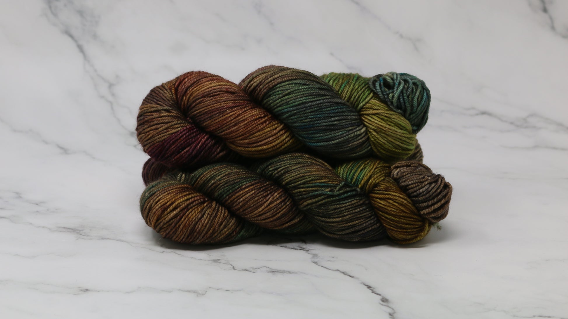 3 skeins of hand-dyed yarn in earthy, variegated shades of green, brown, and burgundy, displayed on a marble surface and background.