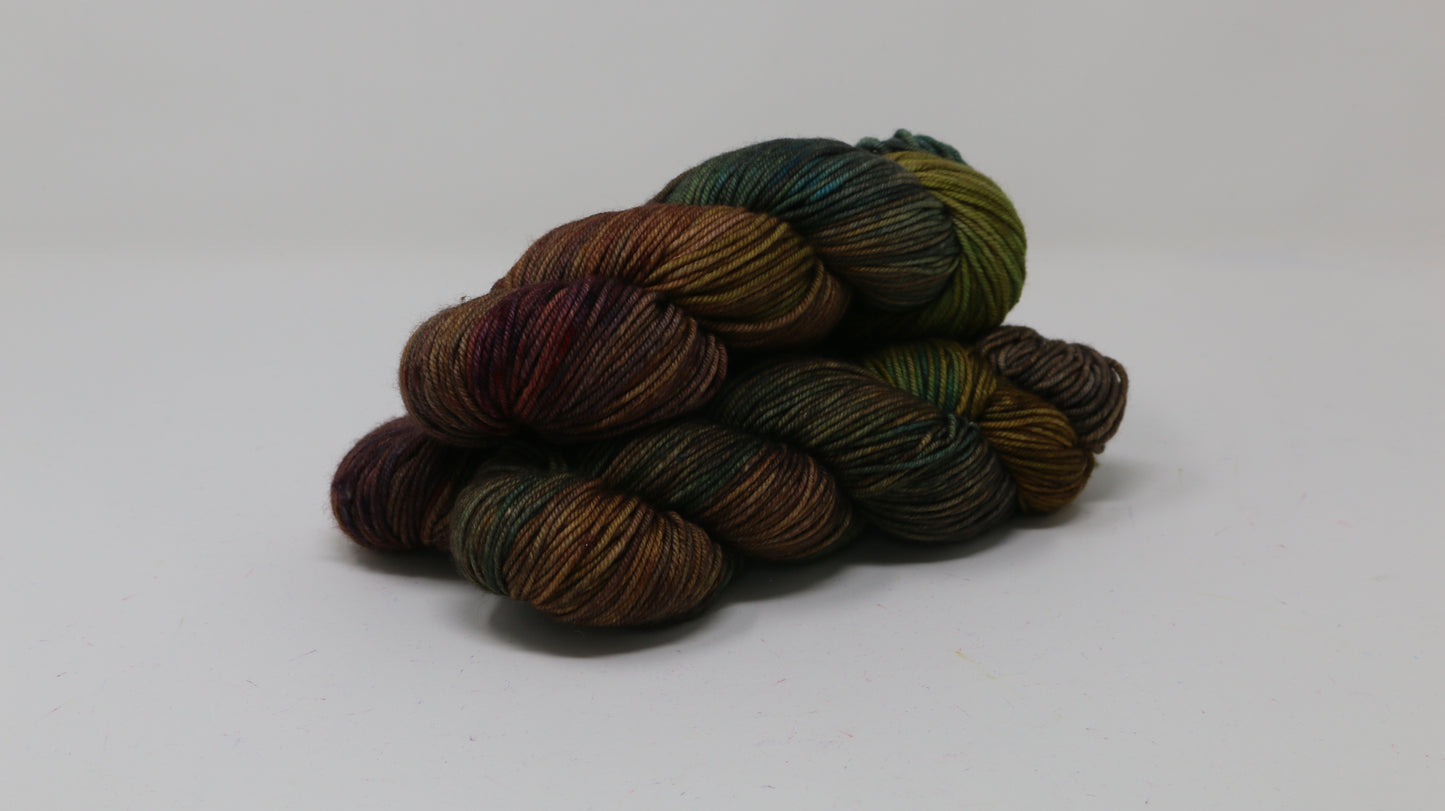 3 skeins of hand-dyed yarn in earthy, variegated shades of green, brown, and burgundy, displayed on a white surface and background.