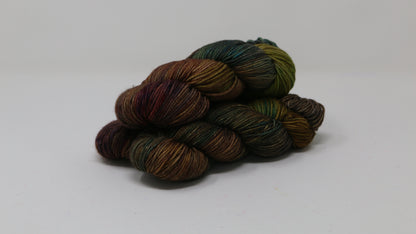 3 skeins of hand-dyed yarn in earthy, variegated shades of green, brown, and burgundy, displayed on a white surface and background.