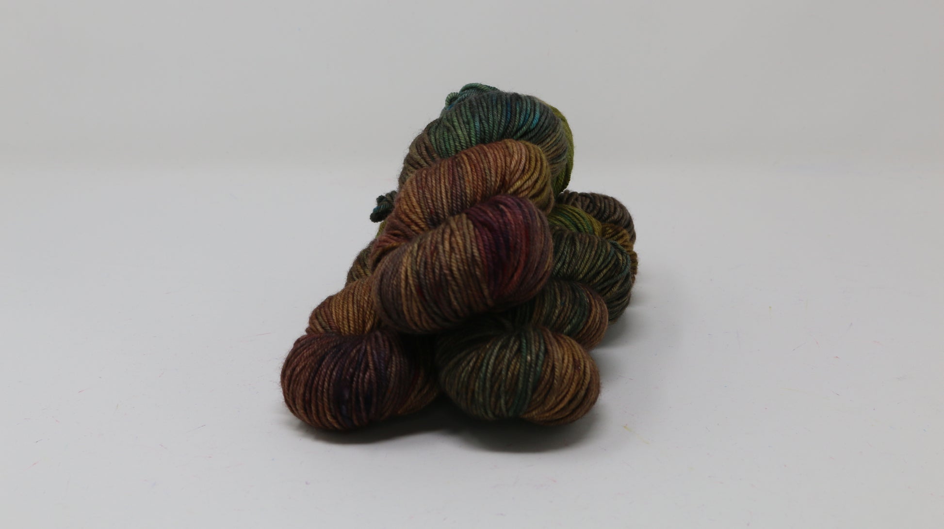 3 skeins of hand-dyed yarn in earthy, variegated shades of green, brown, and burgundy, displayed on a white surface and background.