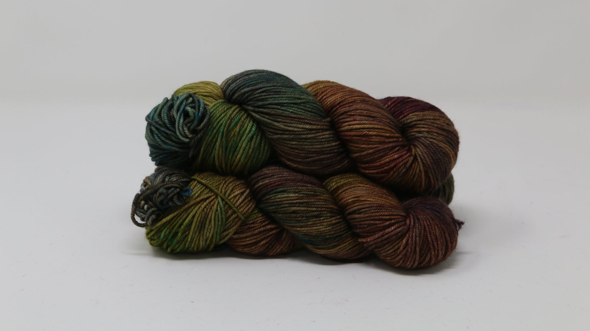3 skeins of hand-dyed yarn in earthy, variegated shades of green, brown, and burgundy, displayed on a white surface and background.