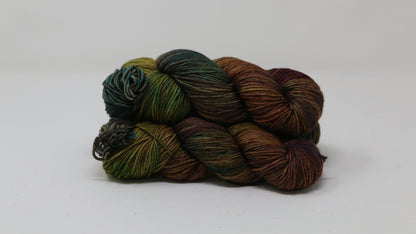 3 skeins of hand-dyed yarn in earthy, variegated shades of green, brown, and burgundy, displayed on a white surface and background.