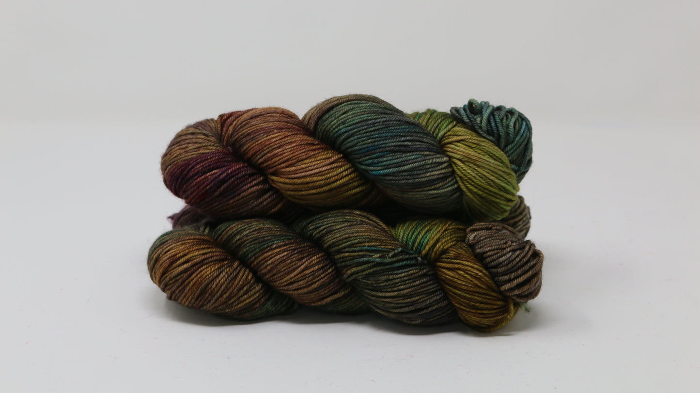 3 skeins of hand-dyed yarn in earthy, variegated shades of green, brown, and burgundy, displayed on a white surface and background.