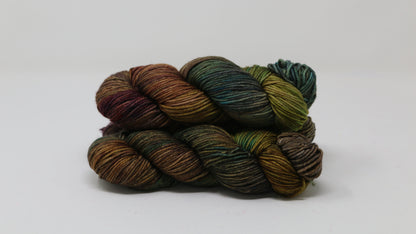 3 skeins of hand-dyed yarn in earthy, variegated shades of green, brown, and burgundy, displayed on a white surface and background.