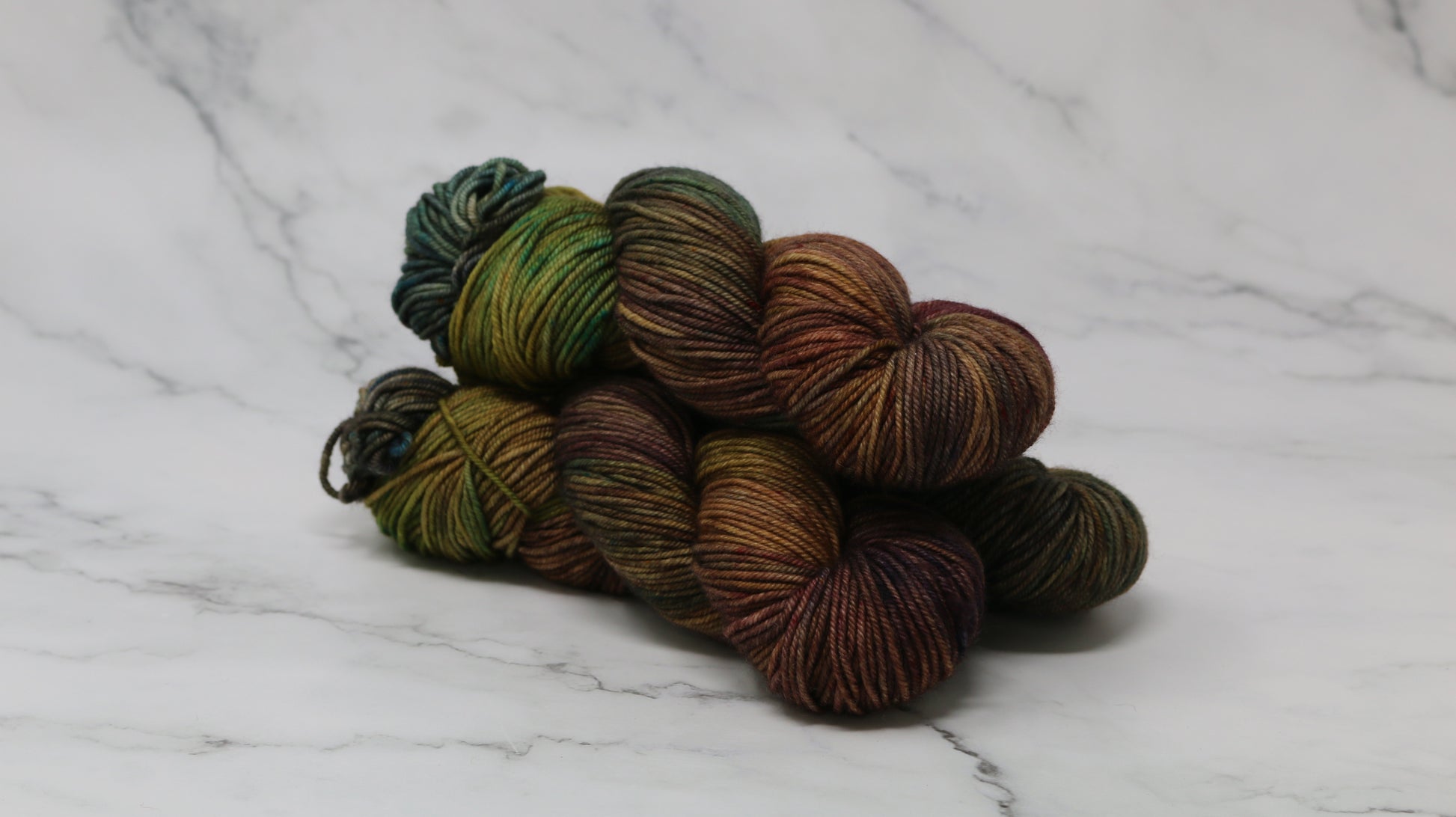 3 skeins of hand-dyed yarn in earthy, variegated shades of green, brown, and burgundy, displayed on a marble surface and background.