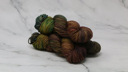 3 skeins of hand-dyed yarn in earthy, variegated shades of green, brown, and burgundy, displayed on a marble surface and background.
