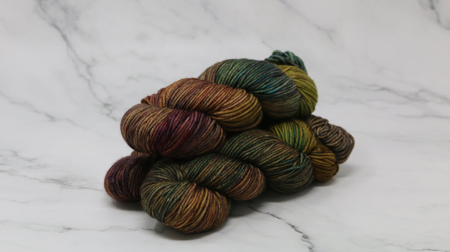 3 skeins of hand-dyed yarn in earthy, variegated shades of green, brown, and burgundy, displayed on a marble surface and background.