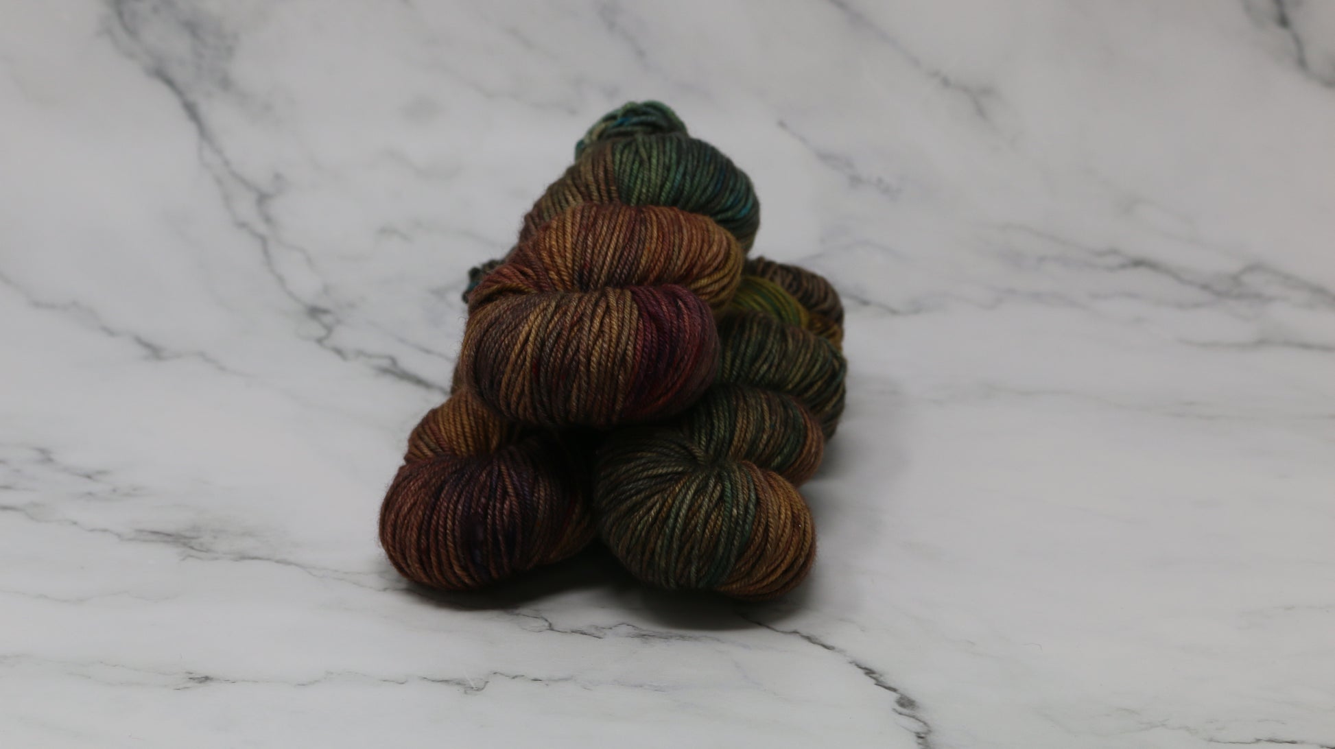 3 skeins of hand-dyed yarn in earthy, variegated shades of green, brown, and burgundy, displayed on a marble surface and background.
