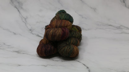 3 skeins of hand-dyed yarn in earthy, variegated shades of green, brown, and burgundy, displayed on a marble surface and background.