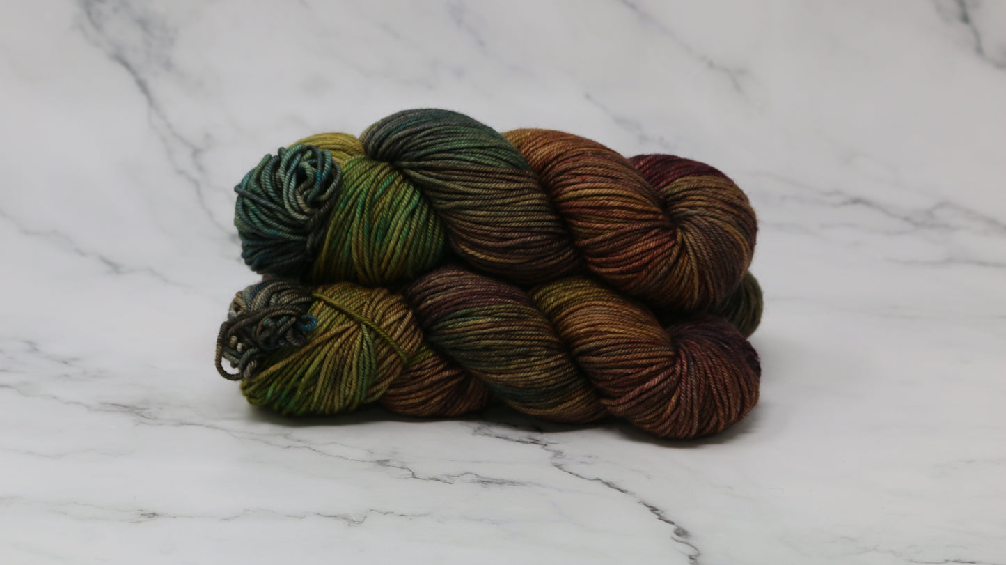 3 skeins of hand-dyed yarn in earthy, variegated shades of green, brown, and burgundy, displayed on a marble surface and background.