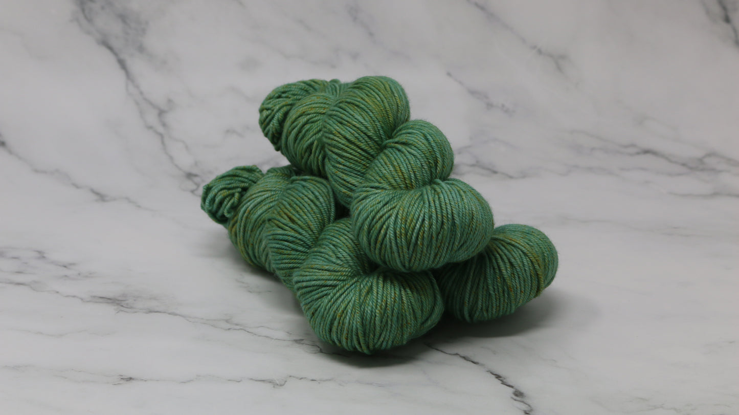 3 skeins of green yarn speckled with gold accents displayed on a white marble surface and background
