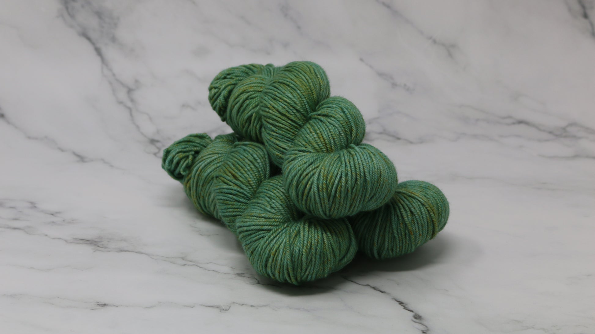 3 skeins of green yarn speckled with gold accents displayed on a white marble surface and background