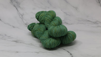 3 skeins of green yarn speckled with gold accents displayed on a white marble surface and background