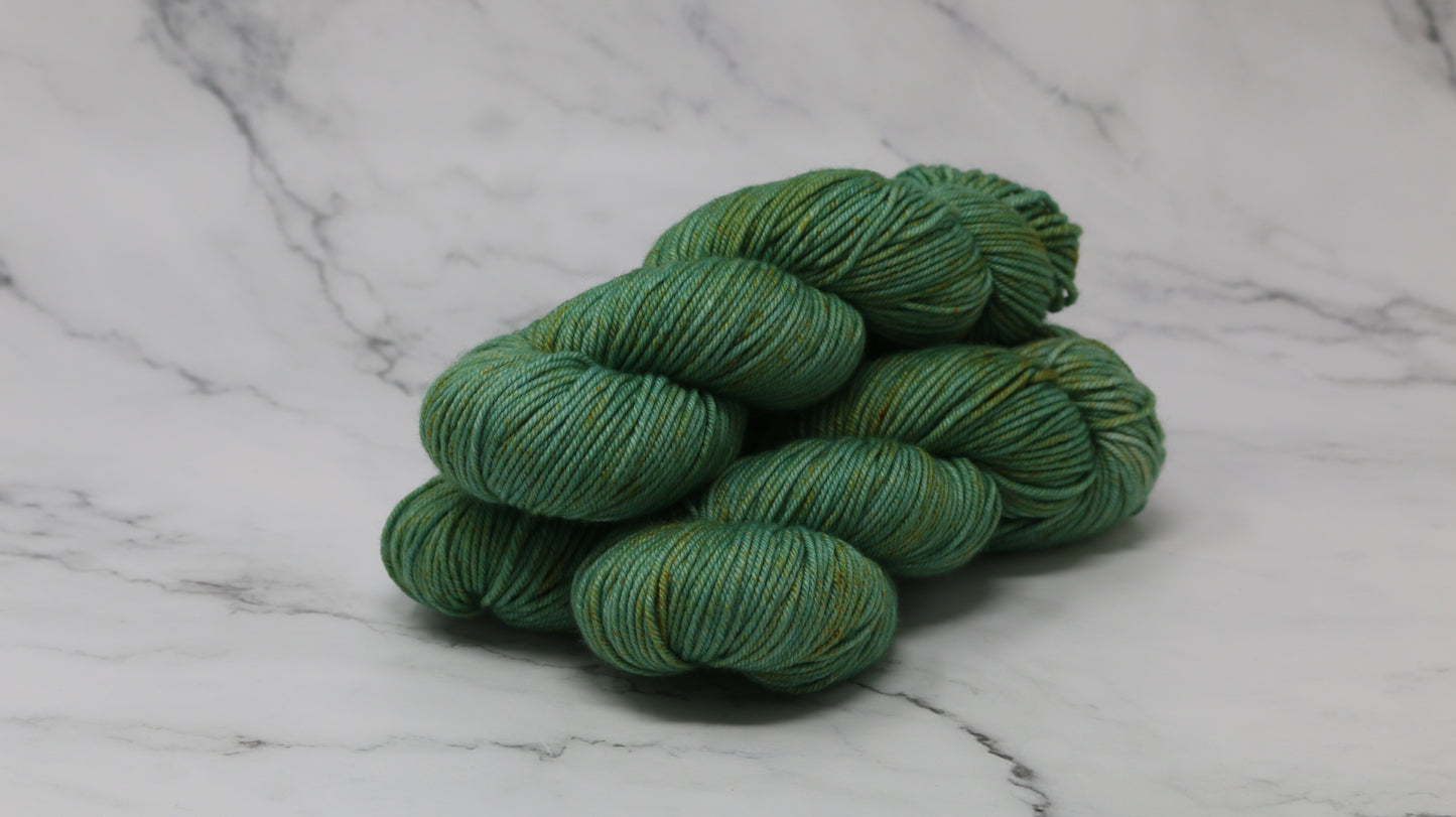 3 skeins of green yarn speckled with gold accents displayed on a white marble surface and background