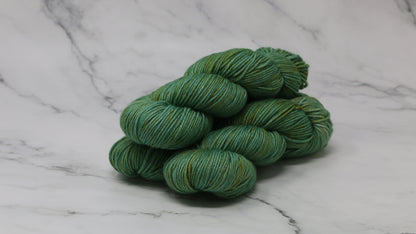 3 skeins of green yarn speckled with gold accents displayed on a white marble surface and background