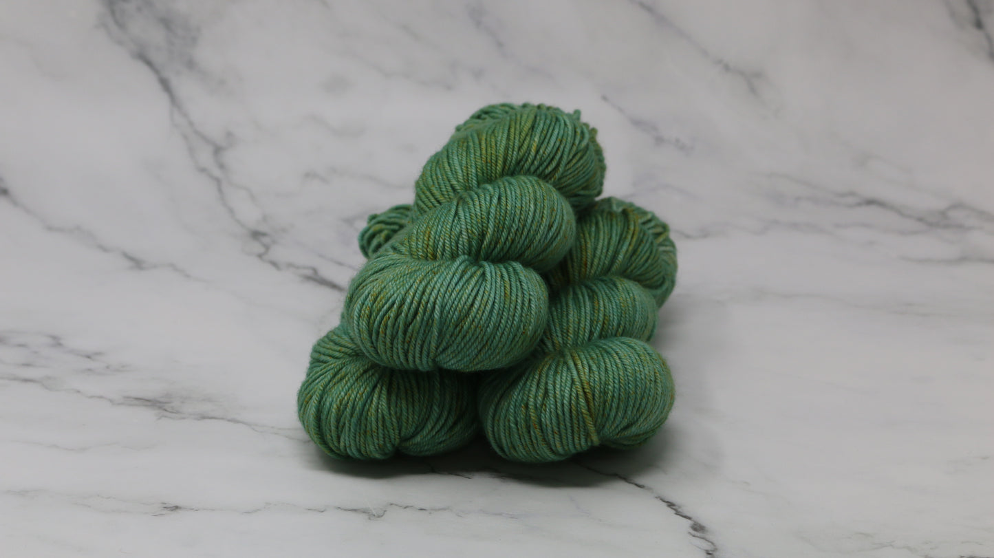 3 skeins of green yarn speckled with gold accents displayed on a white marble surface and background