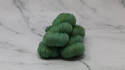 3 skeins of green yarn speckled with gold accents displayed on a white marble surface and background