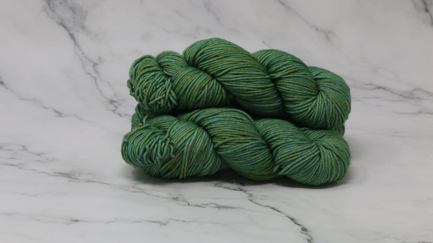 3 skeins of green yarn speckled with gold accents displayed on a white marble surface and background