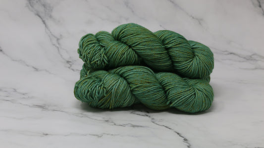 3 skeins of green yarn speckled with gold accents displayed on a white marble surface and background