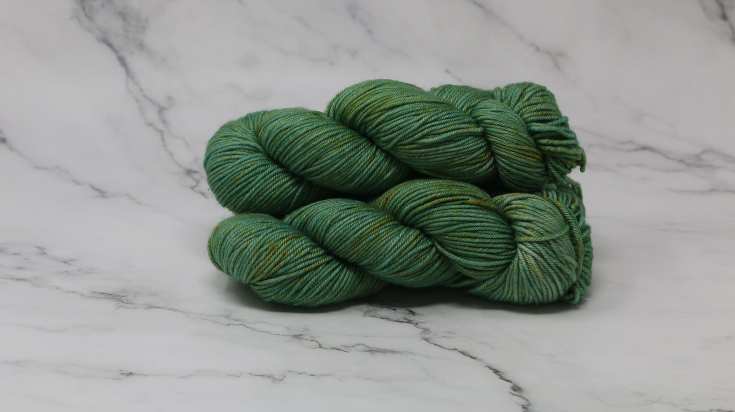 3 skeins of green yarn speckled with gold accents displayed on a white marble surface and background