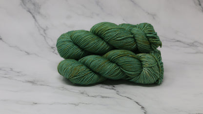 3 skeins of green yarn speckled with gold accents displayed on a white marble surface and background